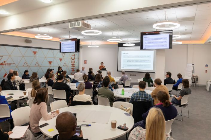 A group of people sit in a seminar room around white tables during a workshop.