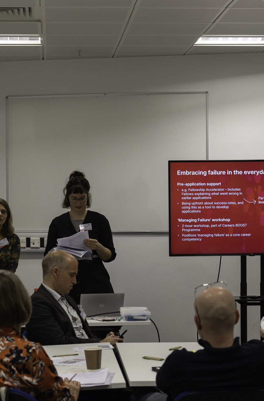 A group of women give a presentation in front of a screen in a seminar room, while people sat at tables listen to them.