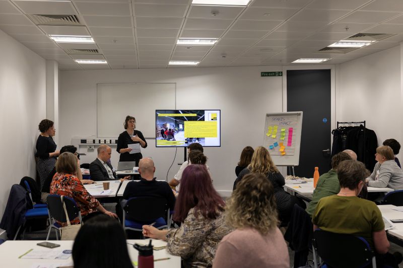 A group of people in a seminar room watching a woman presenting in front of a TV screen.