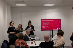 Three women present in front of a screen at the front of a workshop seminar. People sit at tables to watch them,