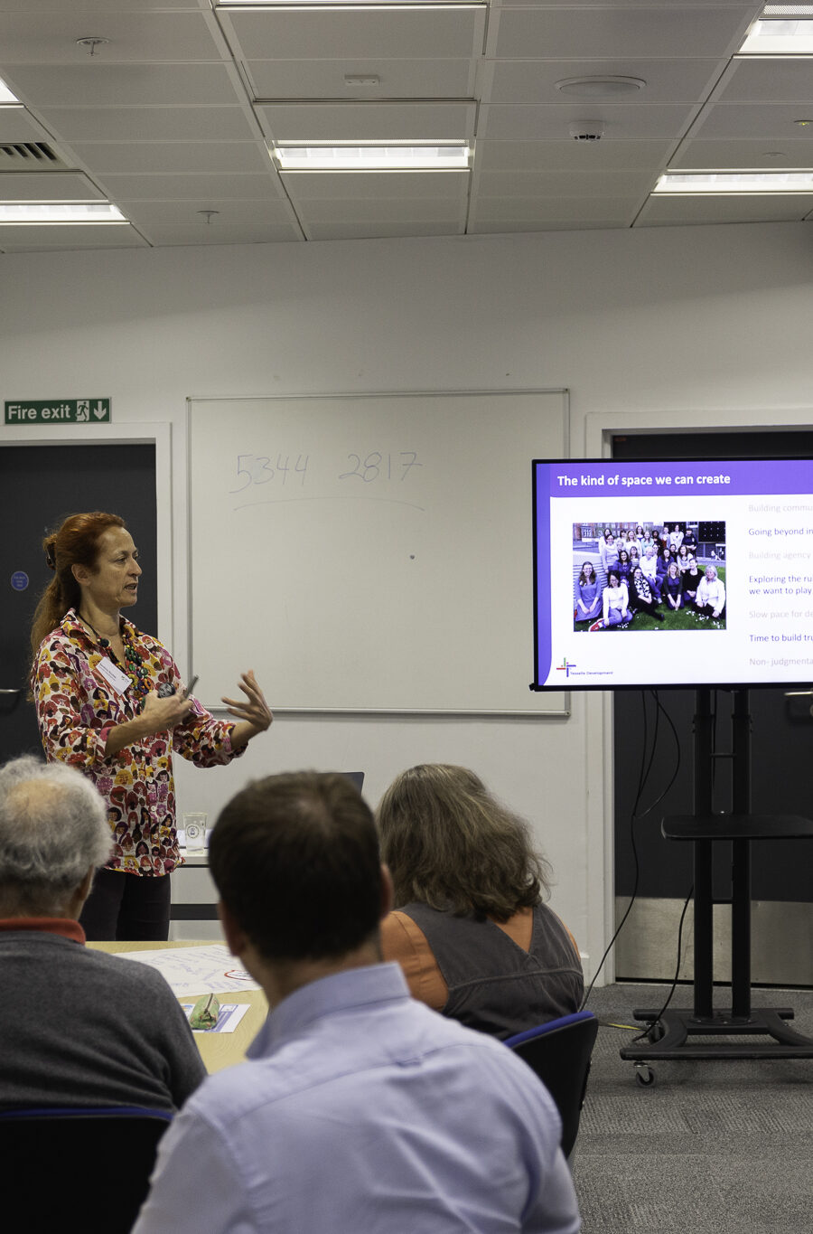 A woman gives a presentation in a seminar room next to a presentation screen.