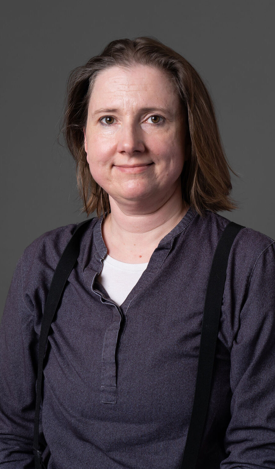 A woman with dark hair wearing a dark grey shirt, in front of a dark grey backdrop.