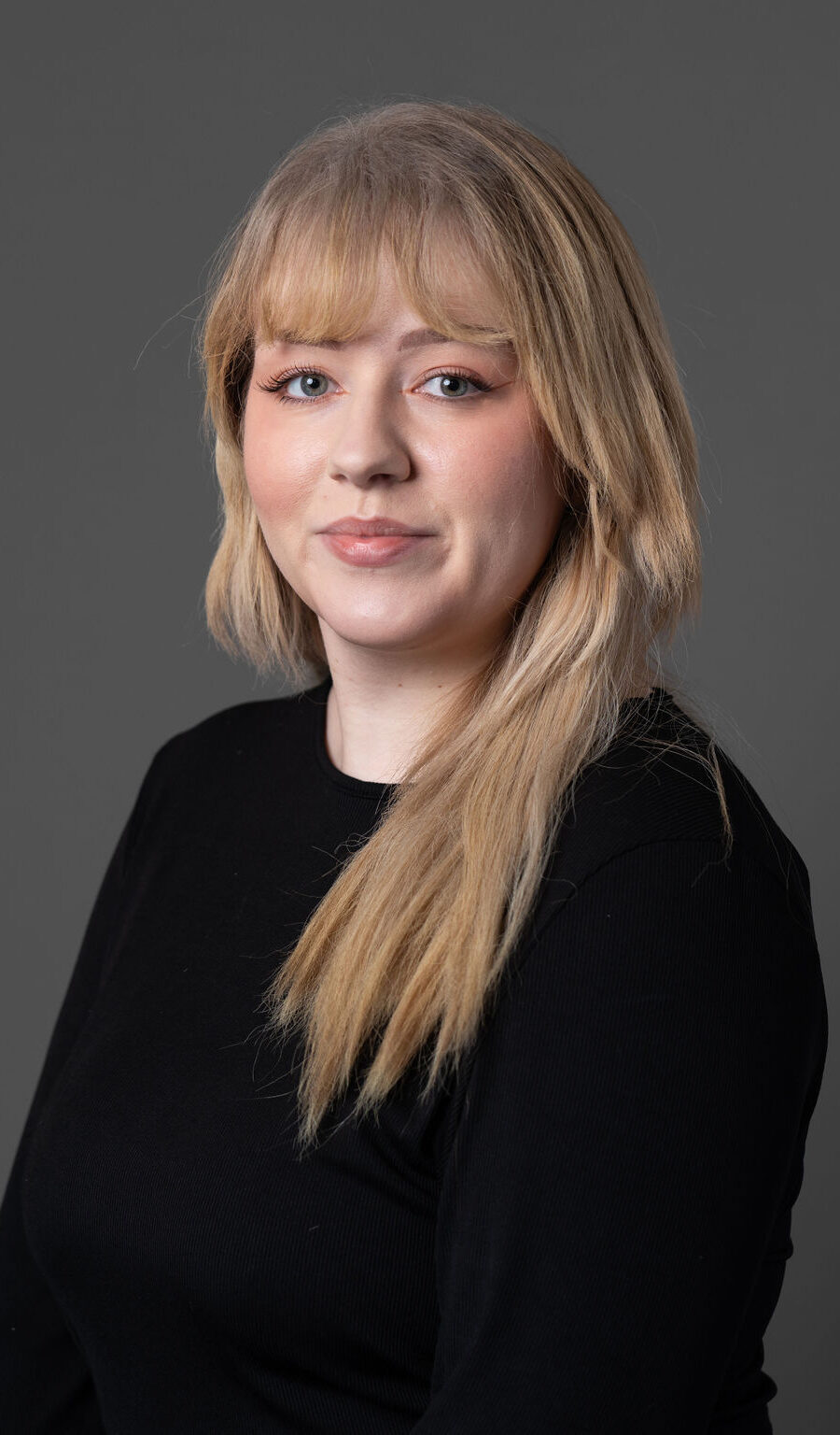 A woman with blonde hair wearing a black top in front of a grey background.