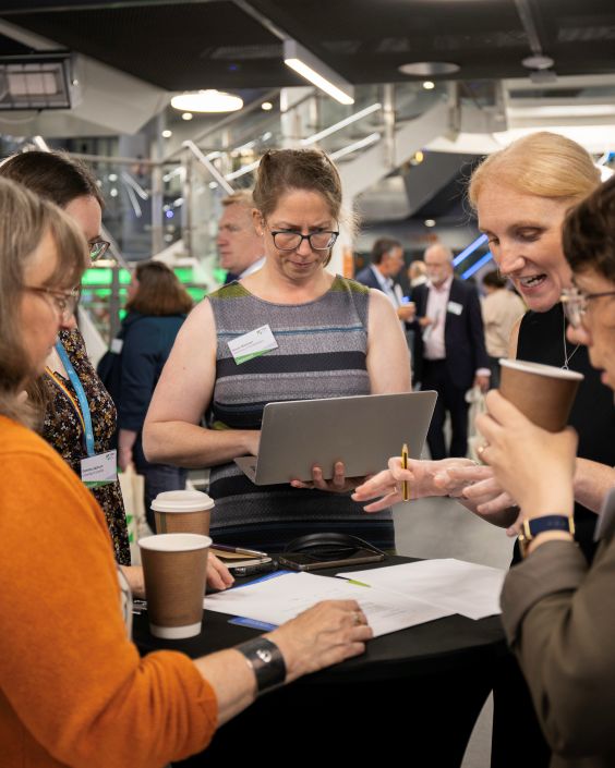 A group of women gather around a table, discussing a paper; one woman is working on a laptop, and all hold coffee cups.