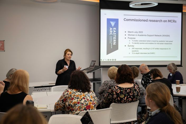 A woman stands at the front of a seminar room and gives a presentation in front of a screen.