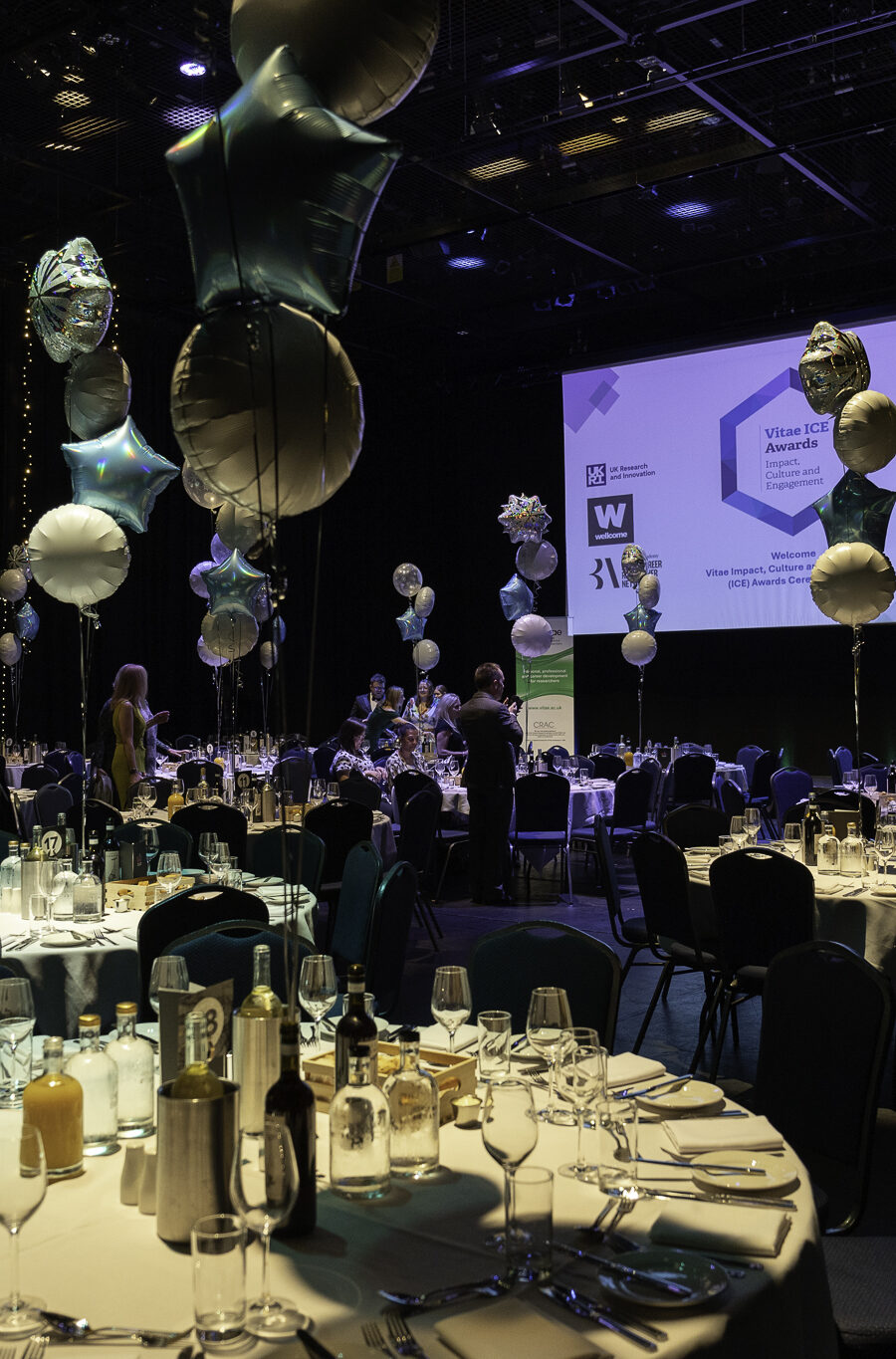 A dinner table at a ceremony, laid out with cutlery and decorated with balloons. A screen is in the background with the Vitae ICE Awards logo.