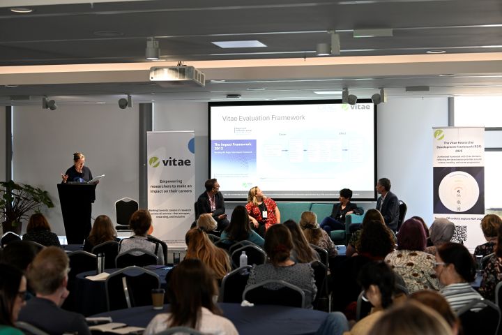 An audience looking at a screen with presenters sat on a stage with another presenter at a podium. Image from #VitaeCon2025 by Andrew Callaghan.