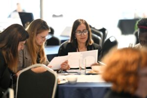 A group of women read pieces of paper while sat at a table.  Image from #VitaeCon2025 by Andrew Callaghan.