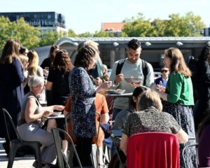 A group of people stand and sit outside on a balcony, talking and eating.  Image from #VitaeCon2025 by Andrew Callaghan.