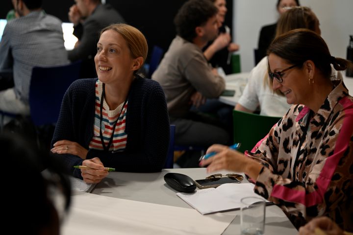 A pair of women smiling and sitting at a table while a workshop happens around them. Image from #VitaeCon2025 by Andrew Callaghan.