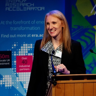 Dr Katy Mahoney standing at a podium with a blue and green background behind her.