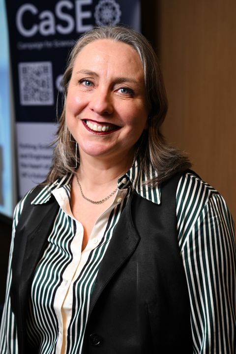 Dr Alicia Greated - a woman smiling at a camera with dark shoulder length hair wearing a striped shirt and dark waistcoat.