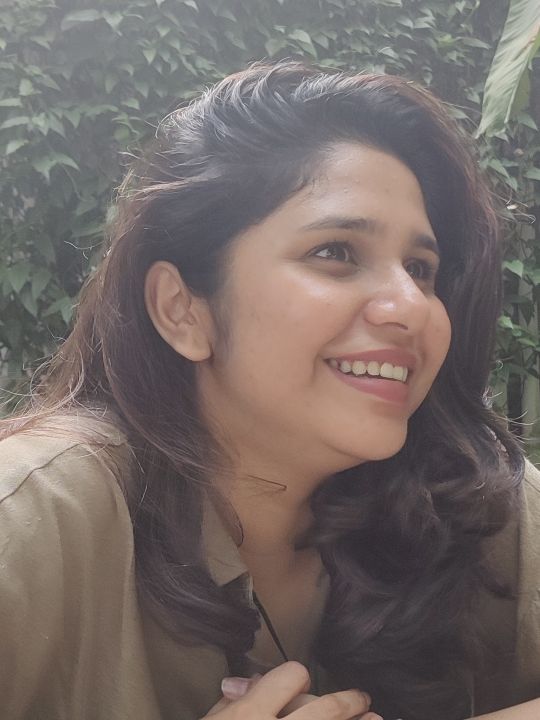A brown woman with dark hair looks off of camera with greenery behind her.