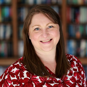 A white woman with dark hair wearing a red patterned top standing in front of a bookshelf.