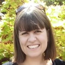 A white woman with brown hair and a fringe stands in front of some trees.