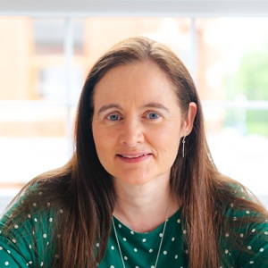 A white woman with dark brown hair wearing a green top in front of a light background.