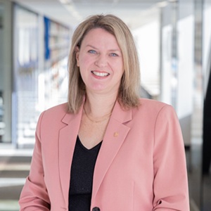 A white woman with blonde hair wearing a pink jacket looking at the camera, standing in front of windows and a staircase.