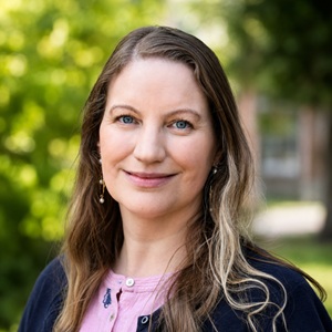 A white woman with long brown hair stands outside with trees behind her.