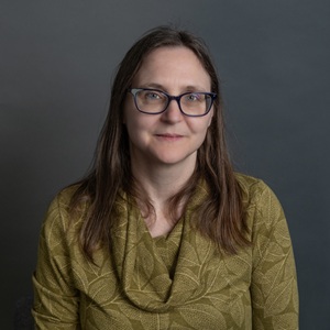 A white woman wearing glasses and a green leaf patterned top with brown hair sitting in front of a dark grey background.