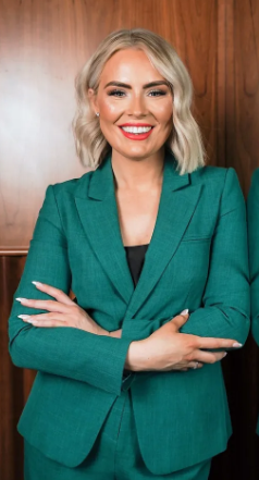 Dr Patricia O'Lynn - A woman stands with folded arms, wearing an emerald green suit in front of a wooden panel backdrop, she has blonde hair and is smiling.