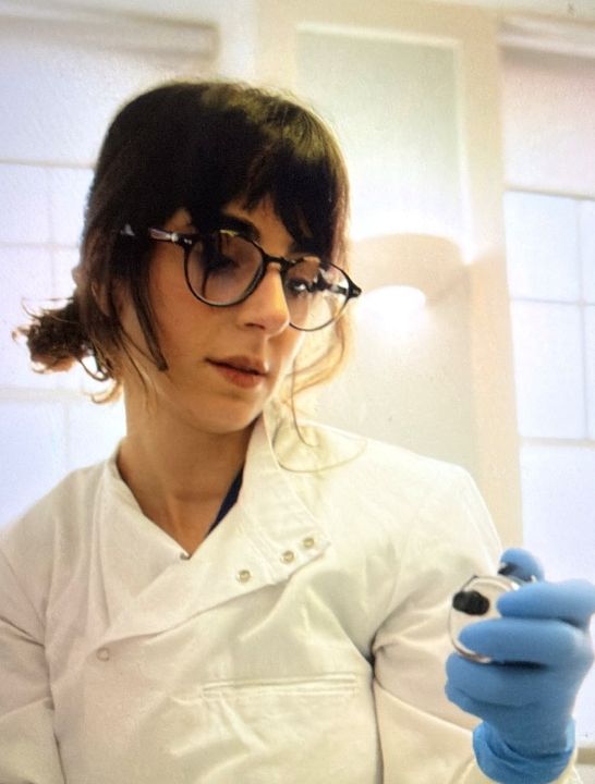 A white woman with dark hair wearing glasses and a white lab coat, holding an instrument while wearing a medical blue glove.