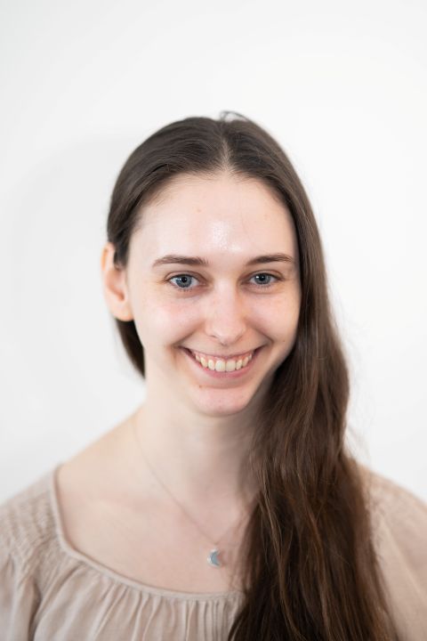 A white woman with long brown hair stands in front of a plain background.