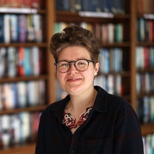 A woman with short cropped hair wears glasses and stands in front of bookshelves.
