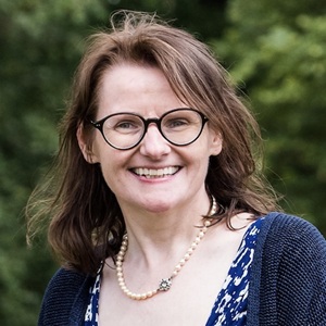 A white woman with dark hair wearing glasses with trees behind her.