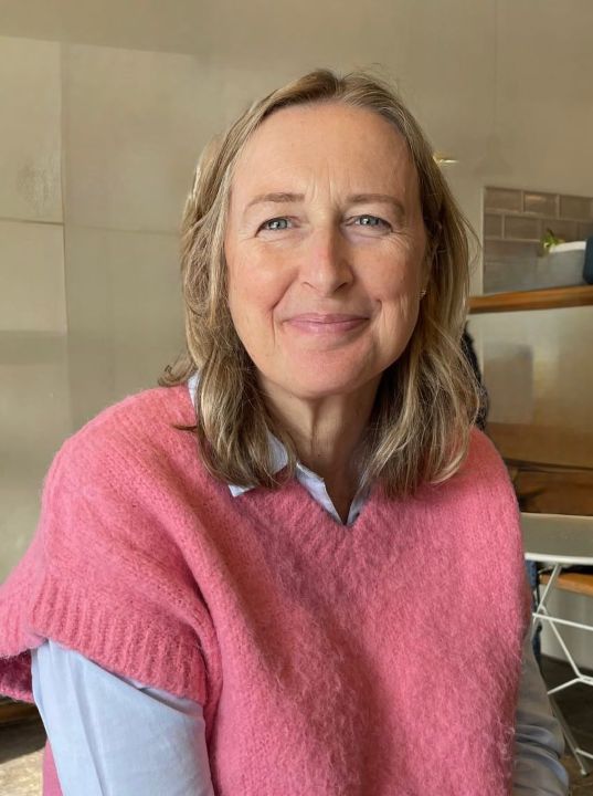 A white woman wearing a pink jumper with short blonde hair.