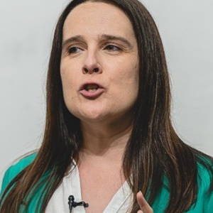 A white woman with dark hair wearing a green jacket, talking with a microphone attached to her top against a plain background.