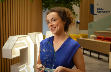 A white woman with brown curly hair holds a glass trophy, sje is wearing a blue top, with the Ulster University logo in top right.