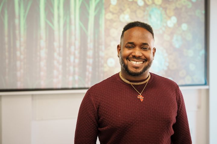 A man of colour with a beard wearing a russet jumper standing in front of a presentation screen.