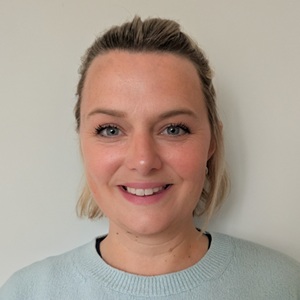 A white woman with short hair stands against a plain background.