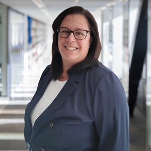 A white woman wearing dark glasses with dark hair, wearing a navy jacket standing in front of a staircase and windows in the background.