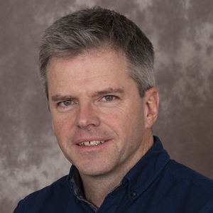 A man wearing a dark shirt in front of a grey dappled background.