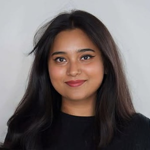 A brown woman with dark brown hair wearing a black top standing in front of a grey background.