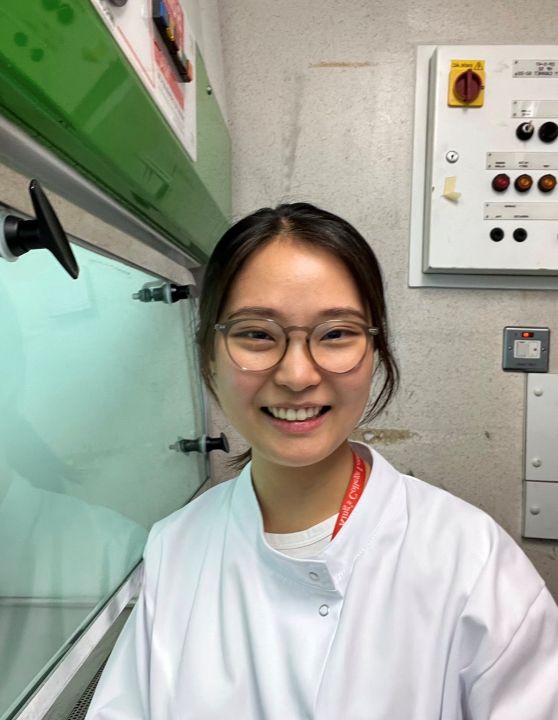 A young woman of Asian descent with glasses wearing a white lab coat, standing next to lab equipment.