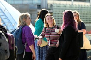 A group of white women in casual clothing stand outside talking with a metal dome in the far left corner behind them. Image from #VitaeCon2025 by Andrew Callaghan.