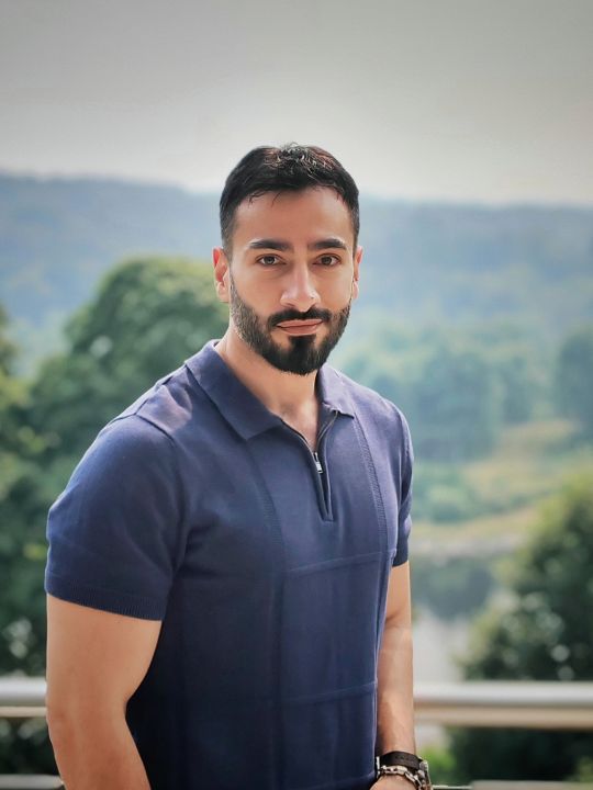 A brown man with a beard wearing a blue top stands in front of a landscape.