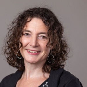 A white woman with brown curly hair smiles at the camera, in front of a grey background.