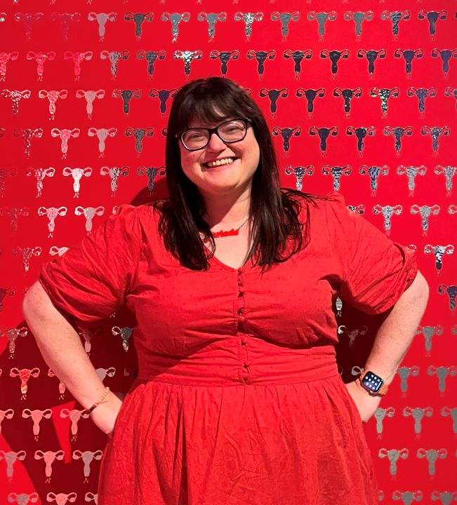A white woman with dark hair and glasses wearing a red dress smiles in front of a red background with the outlines of uteruses around her.