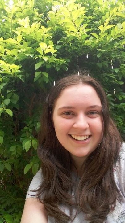 A white woman with long brown hair smiling in front of a green bush.
